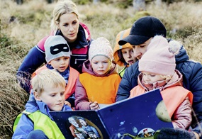 Kinder und Erwachsene sitzen gemeinsam auf einer Wiese, schauen zusammen in ein Buch und lesen miteinander.