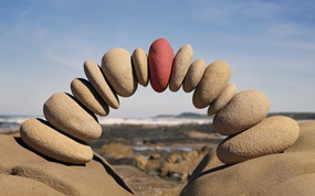 Am Strand werden einzelne Steine zu einem Bogen aufgestapelt, wobei der mittlere Stein rot ist.