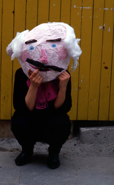 Eine erwachsene Person hält eine Maske aus Kleisterpapier vor ihr Gesicht, die weiße Haare hat.