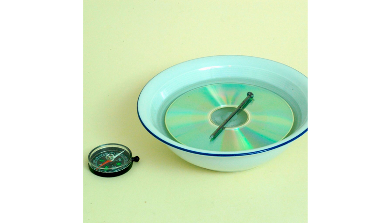 In einer Schale mit Wasser befindet sich eine CD, darüber schwimmt ein Nagel und neben der Schale liegt ein Kompass.