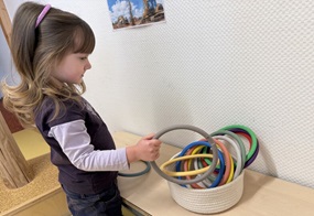 Bea steht vor einem Korb mit bunten Aktivitätsringen und greift zu. 