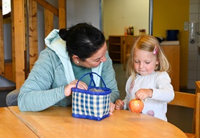 Eine Erzieherin und ein Kind sitzen am Tisch und holen etwas aus einem Korb.