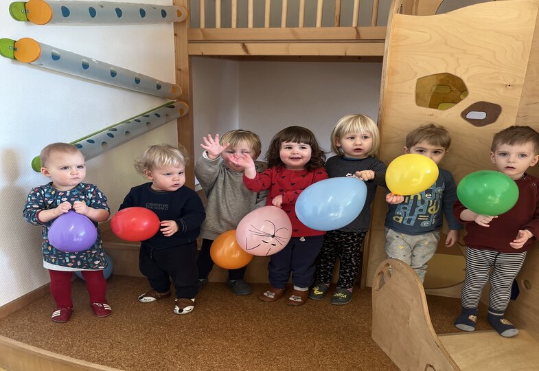 Sieben Kinder stehen in einer Reihe, jedes hält einen bunten Luftballon in der Hand, zwei von ihnen winken.