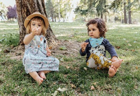 Zwei Kinder sitzen nebeneinander auf einer Wiese, im Hintergrund stehen Bäume.
