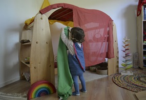 Mit bunten Tüchern baut ein Kind ein Fantasiehaus, indem es die Tücher auf ein Regal- oder Klettermöbelstück legt.