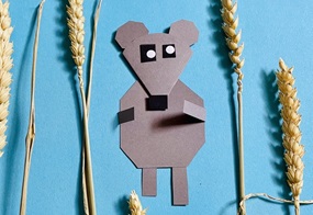 Auf blauem Tonpapier ist eine Maus aus grauem Tonpapier befestigt, umgeben von Getreidehalmen.