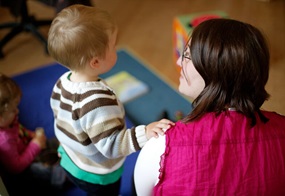 Ein Kleinkind stützt sich mit einer Hand auf der Schulter einer Frau, beide blicken von der Kamera weg.
