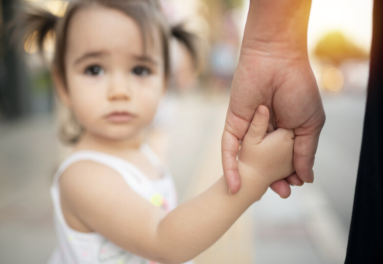 Ein kleines Mädchen blickt bedrückt in die Kamera, im Fokus liegt ihre Hand, die die einer erwachsenen Person hält.