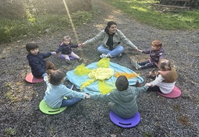 Die Kinder sitzen im Kreis, halten sich gemeinsam an den Händen und in der Mitte liegt eine aus Tüchern gelegte Sonne.