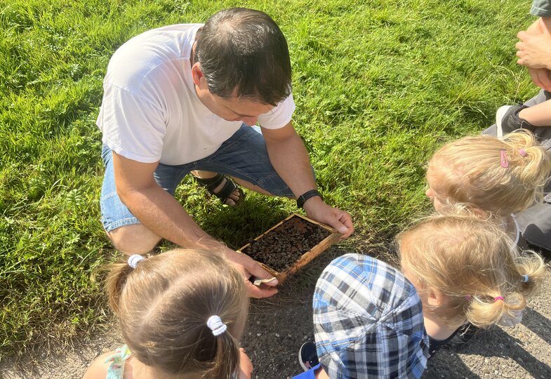 Ein Mann hält ein Wabenrähmchen mit Bienen in der Hand, und rundherum schauen Kinder neugierig zu.