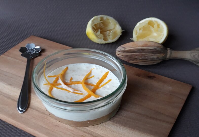 Auf einem Brett steht ein Glas, gefüllt mit Buttermilch-Zitronen-Törtchen, und im Hintergrund liegen die ausgepressten Zitronen.