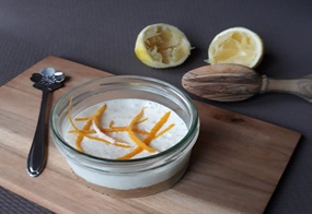 Auf einem Brett steht ein Glas, gefüllt mit Buttermilch-Zitronen-Törtchen, und im Hintergrund liegen die ausgepressten Zitronen.