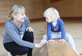 In einer Turnhalle sitzt ein Kind auf einer Bank, daneben hockt eine Frau und schaut das Kind fröhlich an.