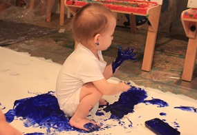 Ein kleines Kind sitzt auf einem großen weißen Tuch, hat blaue Farbe an Hand und Füßen und malt damit auf dieser Fläche.