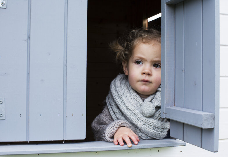 Ein kleines Mädchen mit hellblauem Schal schaut aus einem Fenster mit blauen Fensterläden.