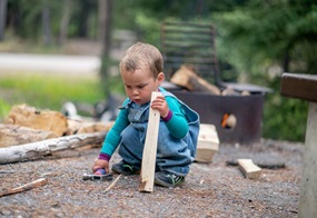 Ein Kleinkind sitzt in der Hocke und hält in der linken Hand ein Stück Holz und in der rechten einen Hammer.