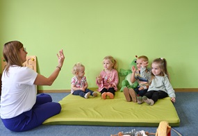 Vor einer grünen Wand auf einer grünen Matte sitzen vier Kinder, und vor ihnen sitzt eine Frau, die ihnen eine Handbewegung vormacht, die sie nachmachen.