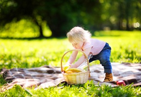 Kleines Mädchen steht auf einer Picknickdecke auf einer Wiese und bückt sich, um etwas aus einem Korb zu holen.
