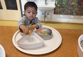 Auf einem runden Tablett stehen mehrere Gegenstände, darunter ein Eisblock mit eingefrorenen Meeresfiguren, und ein Kind füllt mit einer Ballonpipette Flüssigkeit in einen Behälter.