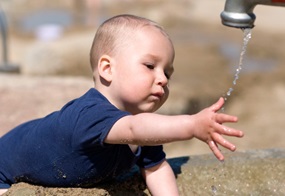 Ein kleiner Junge greift draußen nach Wassertropfen, die aus einem Wasserhahn kommen.