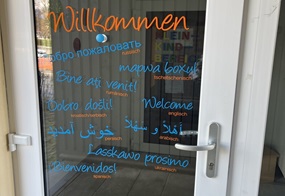 Auf einer Glastür steht mit Glasfarbe in verschiedenen Sprachen „Herzlich willkommen“.