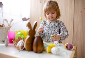 Ein Kind steht an einem Tisch mit ganz viel Osterdeko darauf und hält ebenfalls Osterdeko in der Hand.