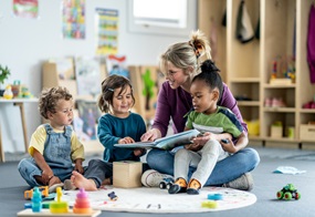 Drei Kinder sitzen zusammen auf dem Boden, während eine Fachkraft ihnen aus einem Buch vorliest.