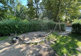 Ein Bereich, der von einer Hecke umgeben ist, zeigt sowohl Gras- als auch Sandflächen, wobei ein Teil mit Naturmaterialien wie Steinen und Ästen abgegrenzt wurde.