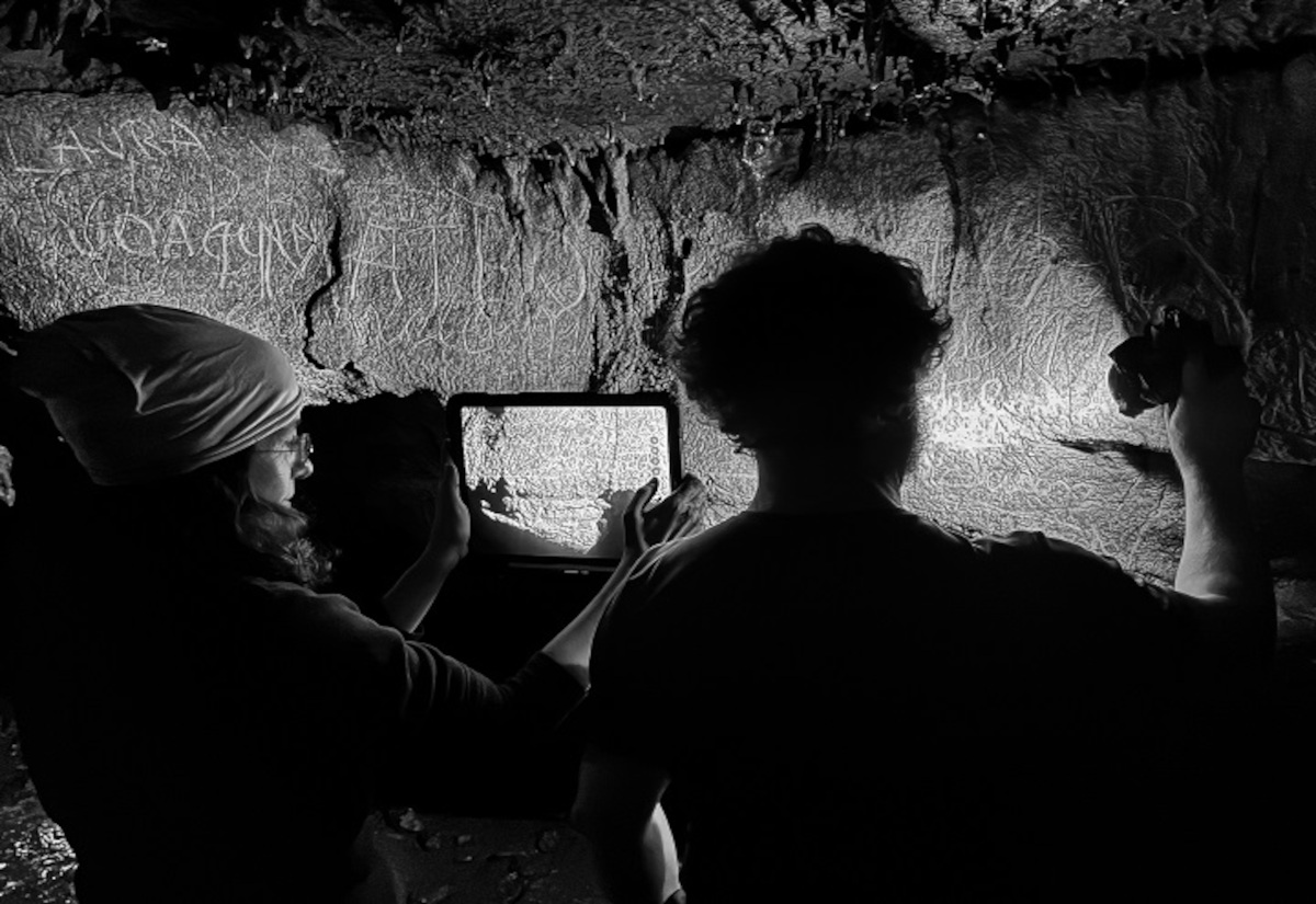 Zwei Personen stehen in einer Höhle vor einer Felswand, die mit zahlreichen eingeritzten Inschriften und Linien bedeckt ist. Eine Person hält ein Tablet oder ähnliches Gerät, das auf die Wand gerichtet ist, während die andere eine Lichtquelle auf die Inschriften richtet. Die Szene ist in schwarz-weiß gehalten, wodurch die Strukturen der Wand und die Umrisse der Personen deutlich hervortreten.