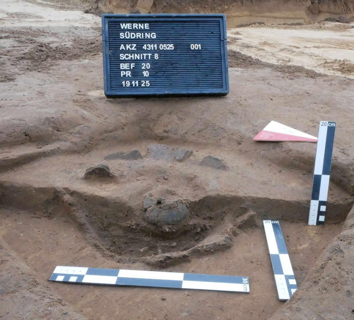 Eine archäologische Ausgrabungsstelle mit freigelegtem Erdreich und mehreren Messlatten. Im Zentrum befindet sich eine runde, Keramik-Struktur. Im Hintergrund steht eine Tafel mit weißer Schrift auf dunklem Grund, die Fundort und weitere Angaben aufführt. Die Messlatten sind weiß mit dunklen Markierungen und Maßangaben. Der Boden ist sandfarben und uneben.