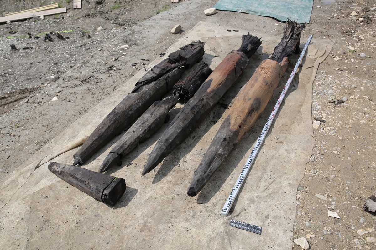 Vier große, spitz zulaufende Holzpfähle liegen auf einer Plane. Ein Maßstab zeigt die Länge der Pfähle an.