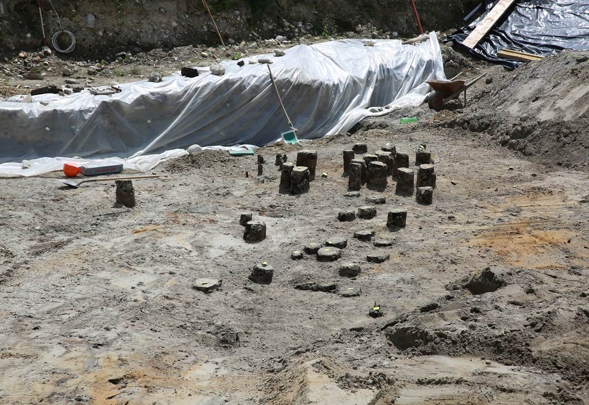 Ausgrabungsstätte mit römischen Brückenfundamenten aus Holz, bestehend aus mehreren runden Stämmen, umgeben von Sand und Folie.