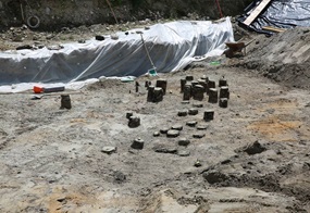 Ausgrabungsstätte mit römischen Brückenfundamenten aus Holz, bestehend aus mehreren runden Stämmen, umgeben von Sand und Folie.