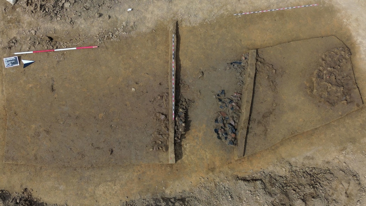 Luftaufnahme der archäologischen Ausgrabungsstätte in Bielefeld-Stieghorst mit zwei rechteckigen Grabungsflächen im Erdreich. Die linke Fläche ist sauber freigelegt, während die rechte Fläche dunkle, unregelmäßige Steinhäufungen zeigt. Am oberen Rand ist ein rot-weißes Maßband und ein kleines Schild zu sehen.