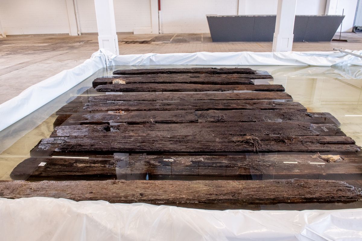 Mehrere alte, dunkle Holzbalken liegen nebeneinander in einer weißen Wanne mit Wasser in einer Halle