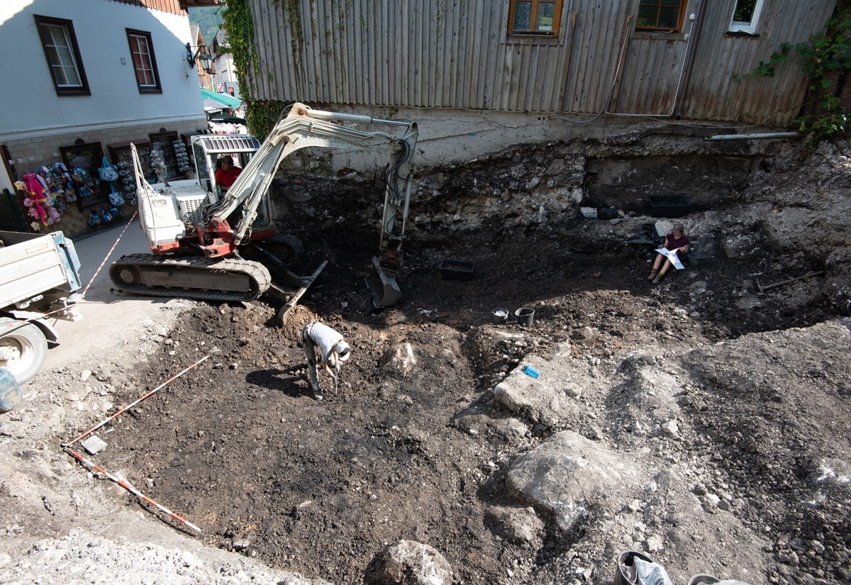 Blick in die Grabungsfläche in Hallstatt Ort, an deren Rand ein Bagger arbeitet. In der Fläche zwei Archäologen bei der Arbeit.