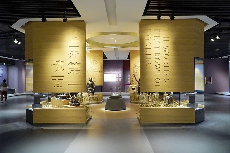 Großer Ausstellungsraum mit goldfarbenen Wänden, die beidseitig mit großflächigen Schriftzeichen versehen sind. In der Mitte steht eine runde Vitrine, umgeben von mehreren Skulpturen, die Menschen bei der Arbeit darstellen. Die Decke ist dunkel und gerippt, der Boden hell und glatt. Im Hintergrund sind weitere Ausstellungsstücke und Wandtafeln zu sehen.