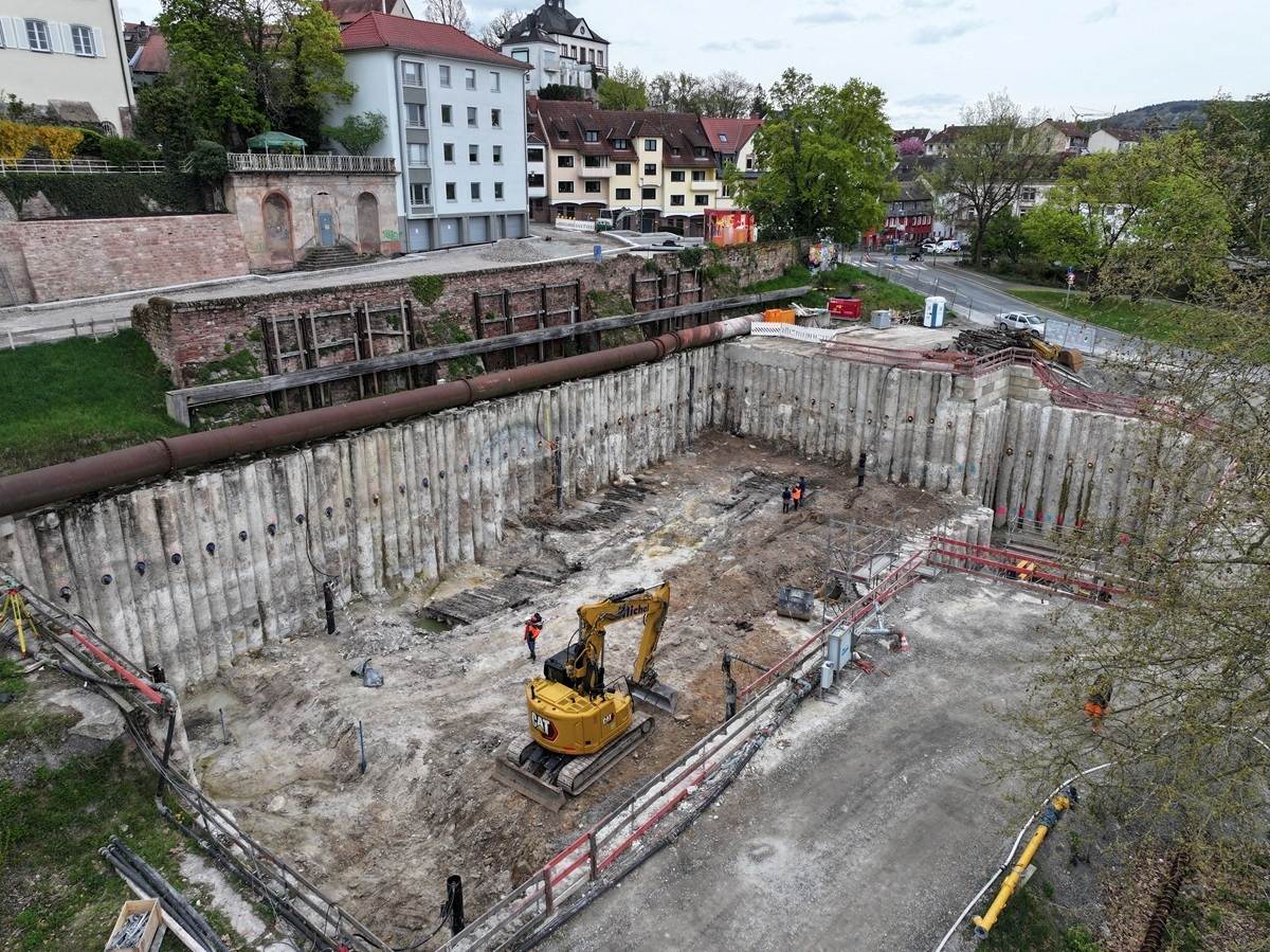 Baugrube mit Bagger und mehreren Personen vor einer historischen Mauer und Wohnhäusern am Flussufer