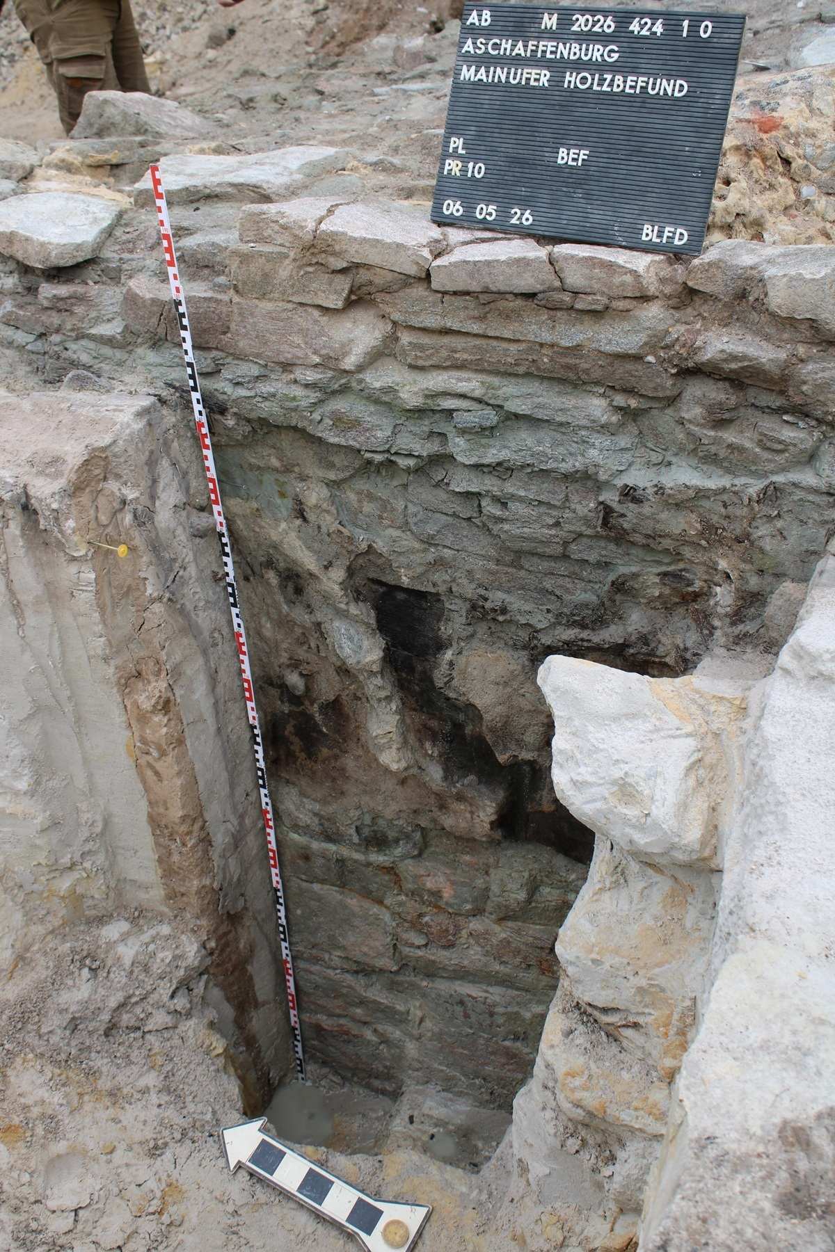 Freigelegter steinerner Bauwerksabschnitt mit Messlatte und beschrifteter Tafel im archäologischen Grabungsbereich