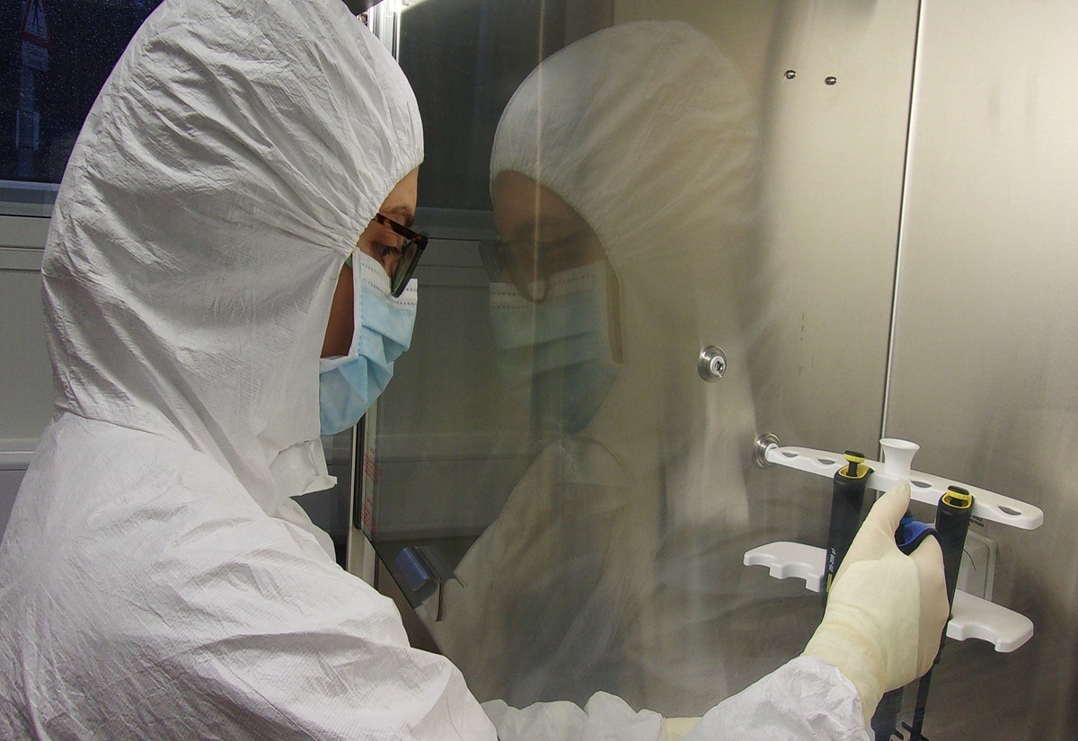 Eine Person trägt einen weißen Ganzkörperschutzanzug, eine Schutzbrille, eine medizinische Maske und Handschuhe. Sie arbeitet in einem Labor an einer sterilen Werkbank und hält eine Pipette in der Hand. Die Umgebung wirkt sauber und technisch, mit reflektierenden Oberflächen und Laborutensilien.