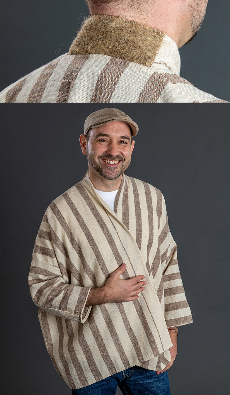 Eine lächelnde Person trägt eine weite Jacke, die sie mit der rechten Hand vorm Bauch geschlossen hält. Die Jacke ist in einem hellbraunen Farbton gehalten. Dunklere Streifen verlaufen parallel und vertikal auf der Jacke.