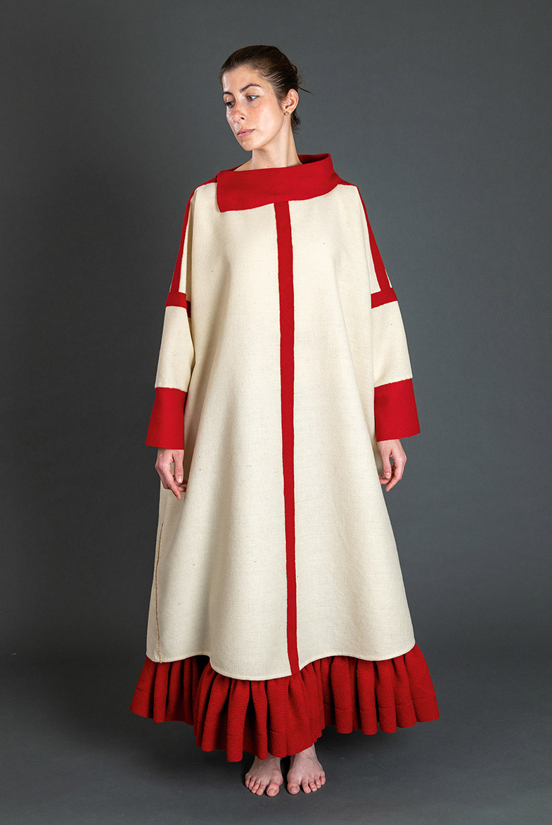 Person steht barfuß in einem langen, cremefarbenen Gewand mit roten Besätzen an Kragen, Ärmeln und Saum vor grauem Hintergrund