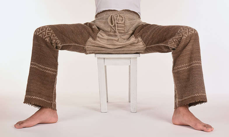 Person sitzt breitbeinig auf einem weißen Hocker und trägt braune, grob gestrickte Hosen mit Mustern an den Oberschenkeln