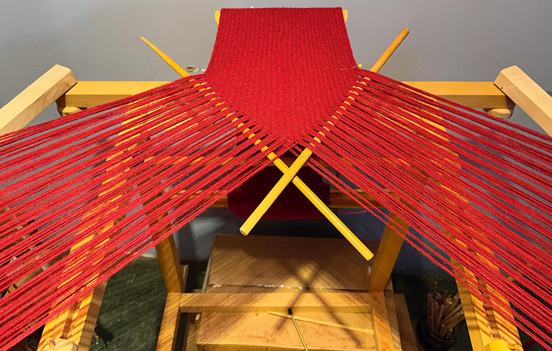 Ein japanischer Flechttisch, in dem zahlreiche rote Fäden fixiert sind, die in der Mitte zu einem gewebten Stück Stoff zusammenlaufen. 