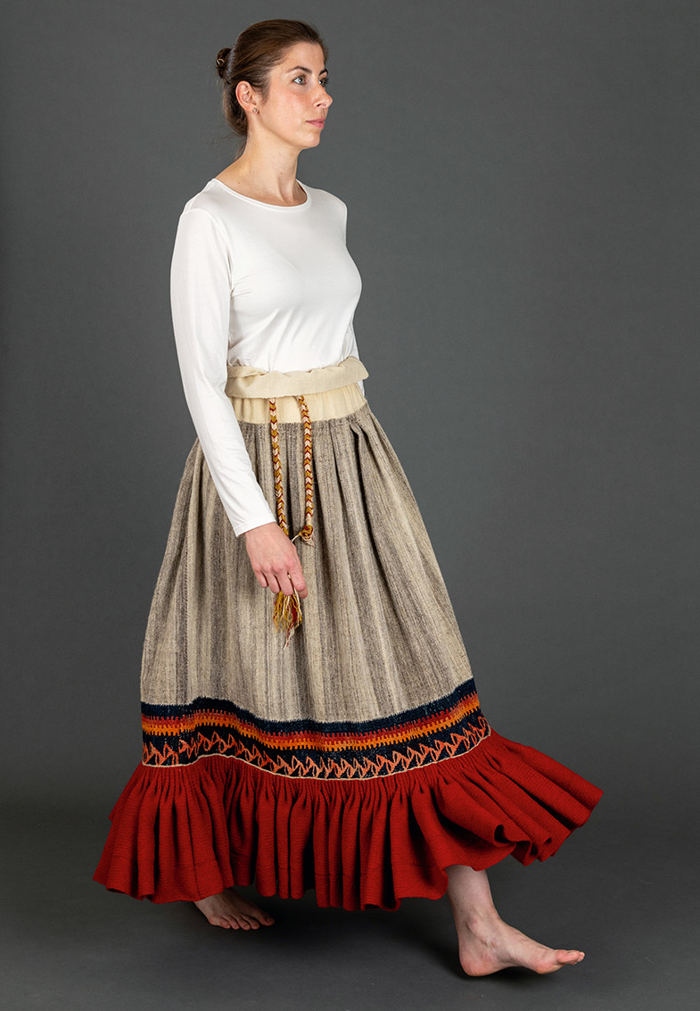 Frau steht barfuß in traditionellem, bodenlangem Rock mit breitem rotem Saum und gemustertem Gürtel sowie weißem Langarmshirt vor grauem Hintergrund