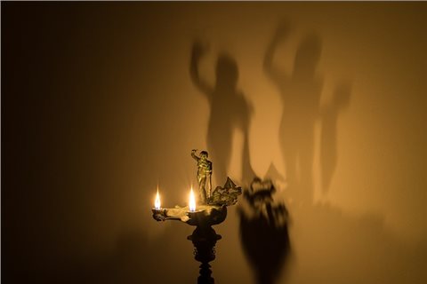 Angezündete Lampe, die mit einem Silen auf dem Deckel geschmückt ist. Durch die doppelte Flamme, wirft das Licht einen doppelten Schatten auf die dahinterliegende Wand. Die Figur wirkt dadurch größer.