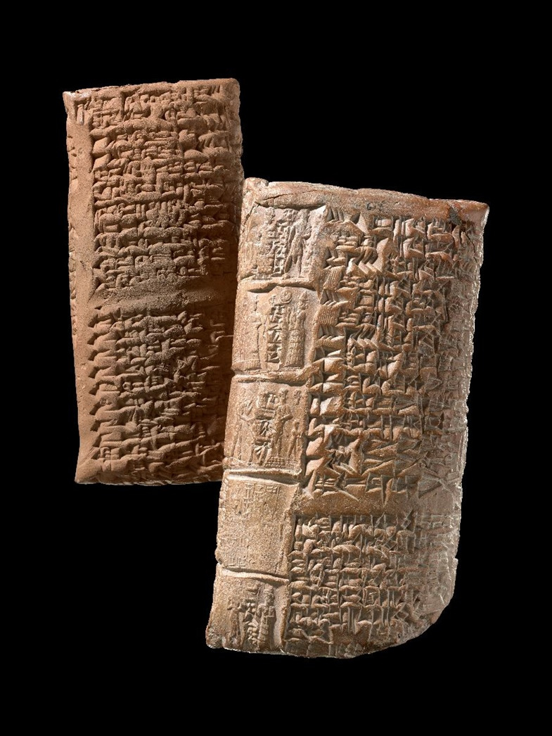 Zwei längliche Tonobjekte mit eingravierten Keilschriftzeichen stehen nebeneinander vor schwarzem Hintergrund. Die Oberflächen sind unregelmäßig und zeigen zahlreiche feine Linien und Vertiefungen, die die Schriftzeichen bilden. Die Farbe der Tonobjekte ist hellbraun.