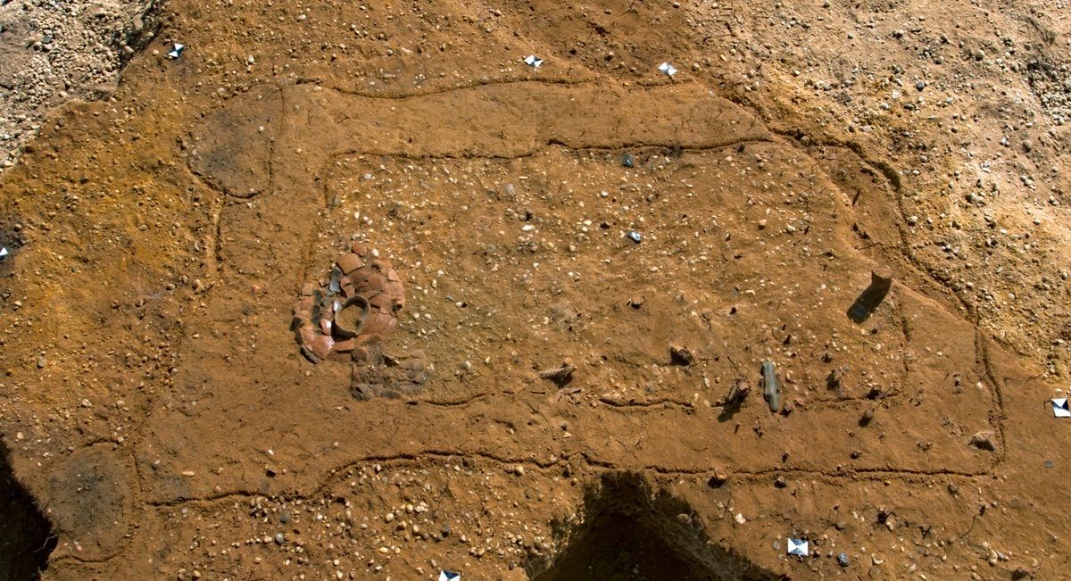 Luftaufnahme einer rechteckigen Ausgrabungsfläche mit klar erkennbaren Umrissen im sandigen Boden. Innerhalb der Fläche sind mehrere kreisförmig angeordnete Tonscherben sichtbar. Kleine weiße Markierungen kennzeichnen an verschiedenen Stellen Funde.