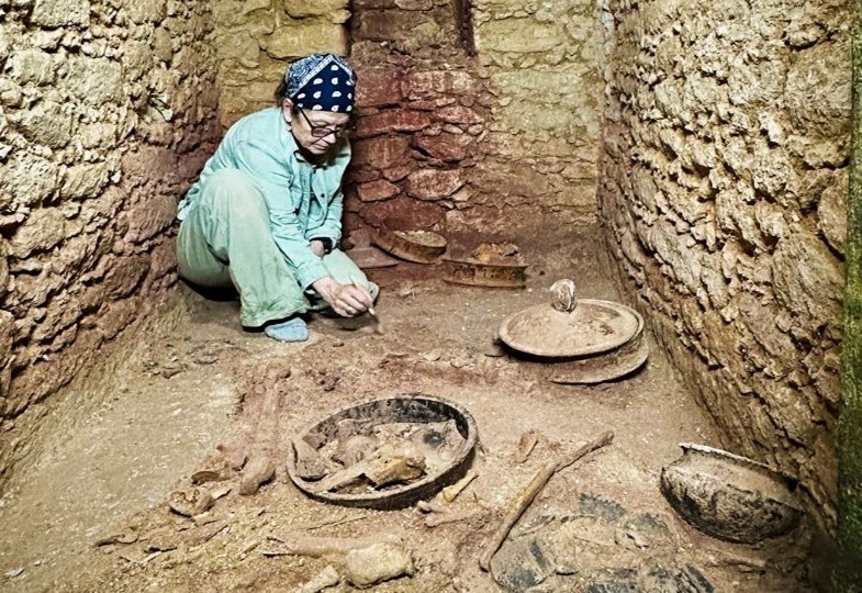 Archäologin Diane Chase in der Grabkammer mit steinernen Wänden untersucht Skelettreste, die neben Keramikschalen auf dem Boden liegen.