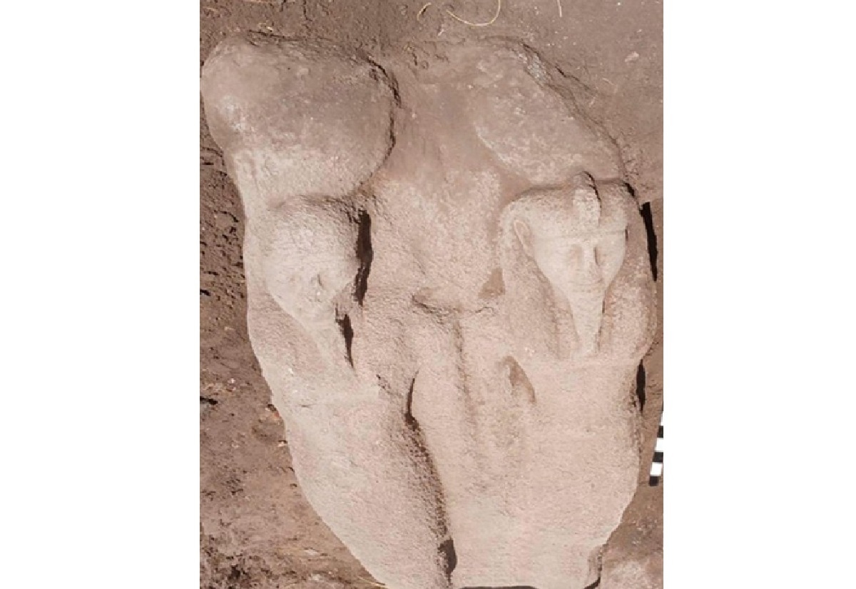 Großer, stark verwitterter Torso einer steinernen Statue mit zwei anliegenden Figuren, die nebeneinander stehen. Die Oberflächen sind uneben und zeigen deutliche Erosionsspuren. Die Köpfe und Körperformen sind nur noch schemenhaft erkennbar. Der Fund liegt auf sandigem Boden, seitlich ist ein Maßstab mit schwarz-weißen Feldern zu sehen.